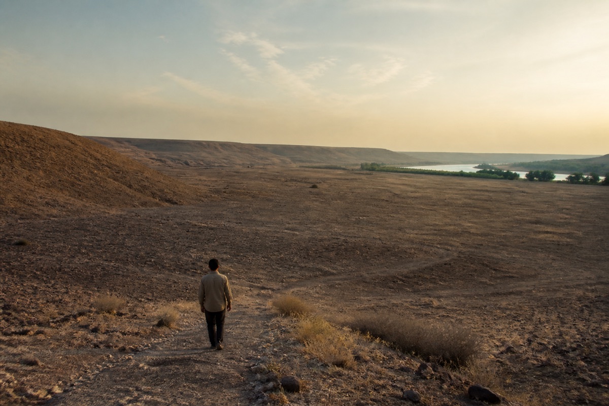 A solitary person crosses a vast dry valley toward a distant river and green tree line.